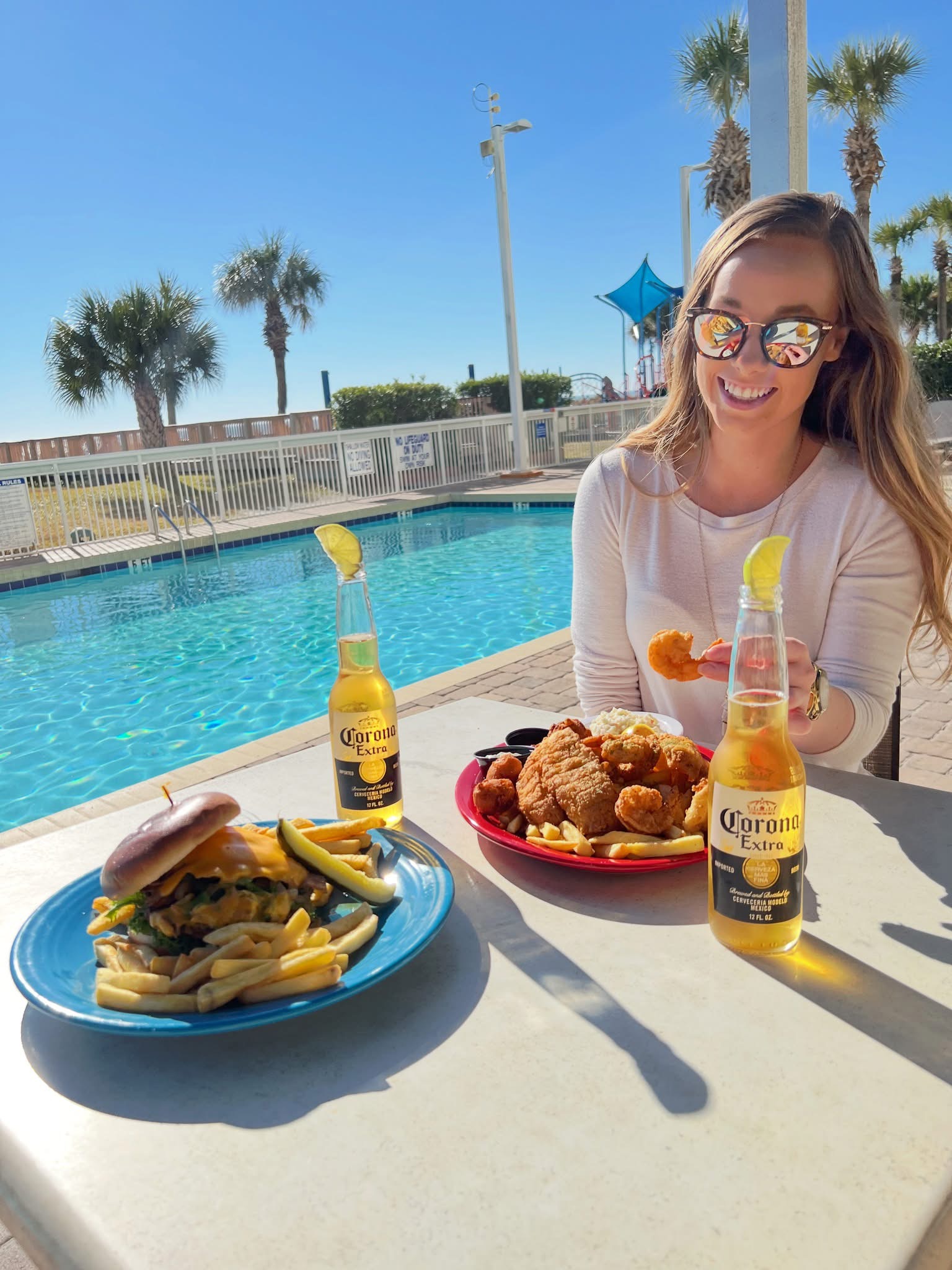 A woman sits at a table by the pool with plates of food and bottles of Corona beer.